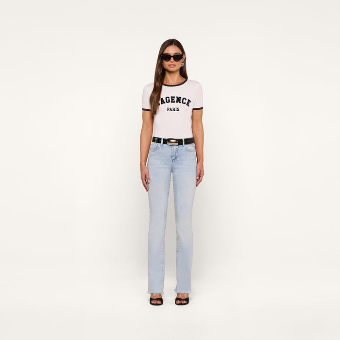 A woman stands against a plain white background wearing sunglasses, light blue jeans with a black belt, black shoes, and the Tommie Crewneck Tee—a relaxed fit Paris tee made from Supima cotton featuring the AGENCE PARIS text.