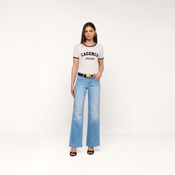 A woman stands against a plain white background wearing a white Tommie Crewneck Tee made from Supima cotton, light blue wide-leg jeans, a black belt, and black high-heeled sandals. She faces forward with her arms relaxed at her sides.