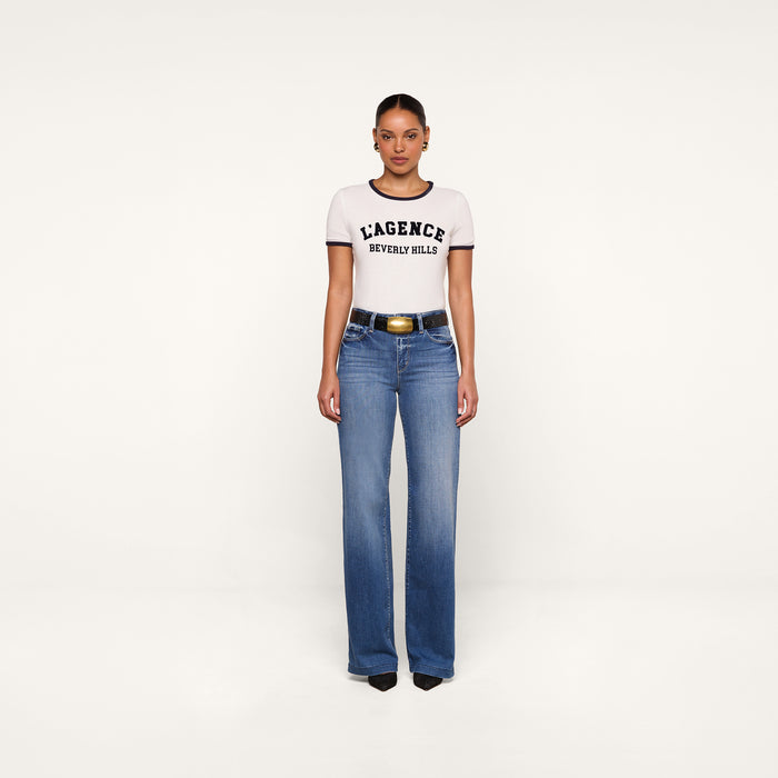 A woman stands facing forward, wearing a relaxed fit white Tommie Crewneck Tee, blue jeans, a black belt with a gold buckle, and black shoes. The background is plain white.