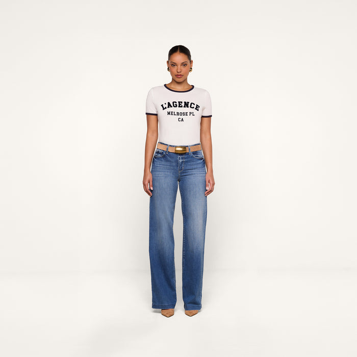 A woman stands facing forward wearing a relaxed fit white Tommie Crewneck Tee, blue high-waisted jeans, a brown belt, and beige heels, against a plain white background.