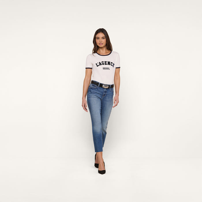 A woman stands against a plain white background wearing a white Tommie Crewneck Tee crafted from Supima cotton, blue jeans, a black belt, and black heels. She faces forward with a neutral expression and arms relaxed by her sides.