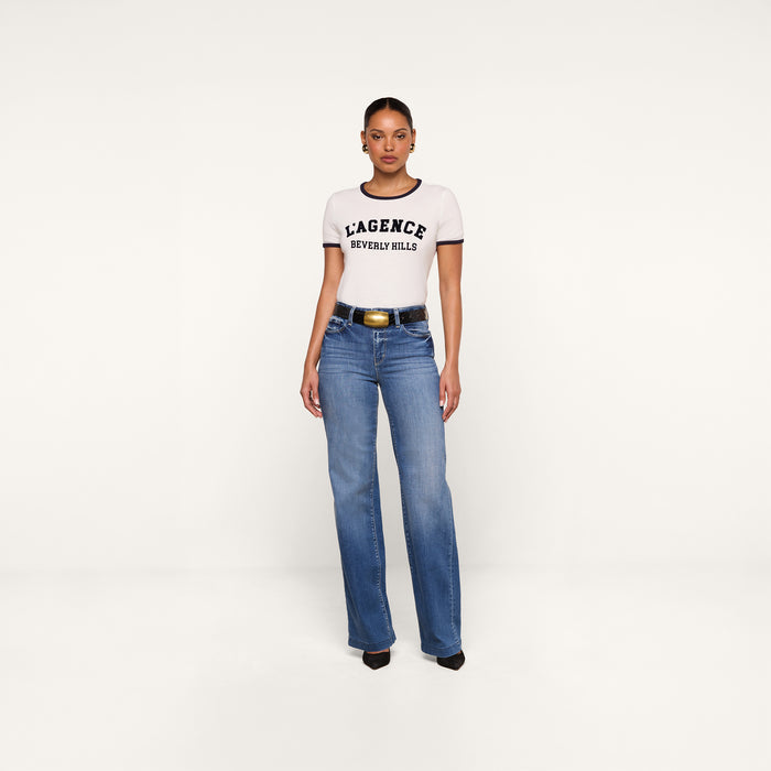 A woman stands against a white background wearing a relaxed fit white Tommie Crewneck Tee made from Supima cotton, blue high-waisted jeans, a black belt with a gold buckle, and black heels.