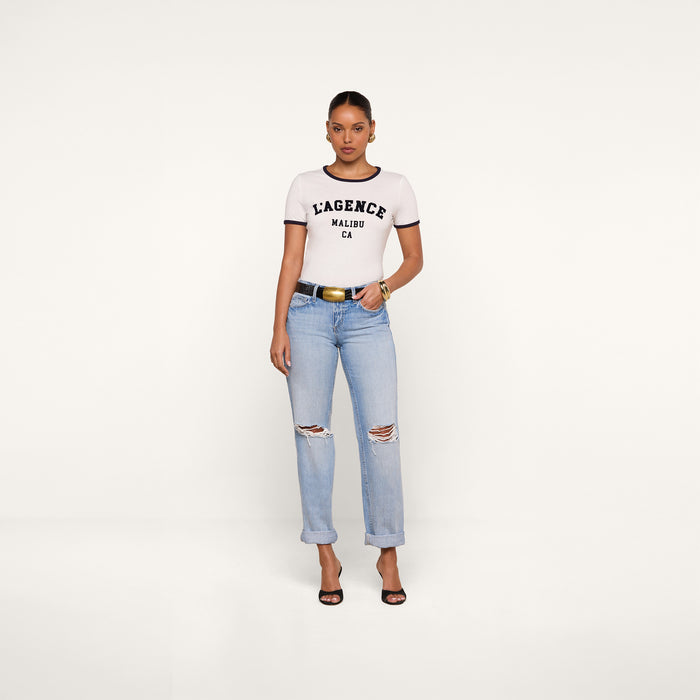 A woman stands against a plain white background, wearing a white Tommie Crewneck Tee crafted from Supima cotton by L’AGENCE, light blue ripped jeans, a black belt, and black high-heeled sandals.