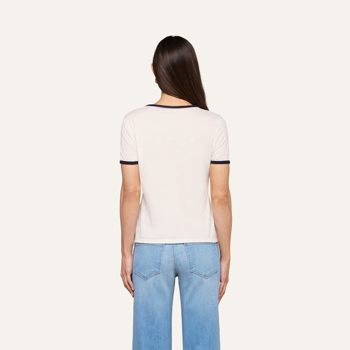 A woman with long brown hair stands facing away, wearing the Tommie Crewneck Tee— a white ringer style with dark trim—paired with light blue jeans against a plain light background.