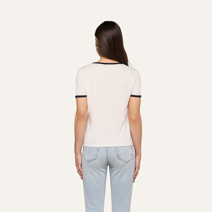 A person with long brown hair, wearing the Tommie Crewneck Tee— a relaxed fit white ringer tee with dark trim—paired with light blue jeans is standing with their back to the camera against a plain background.