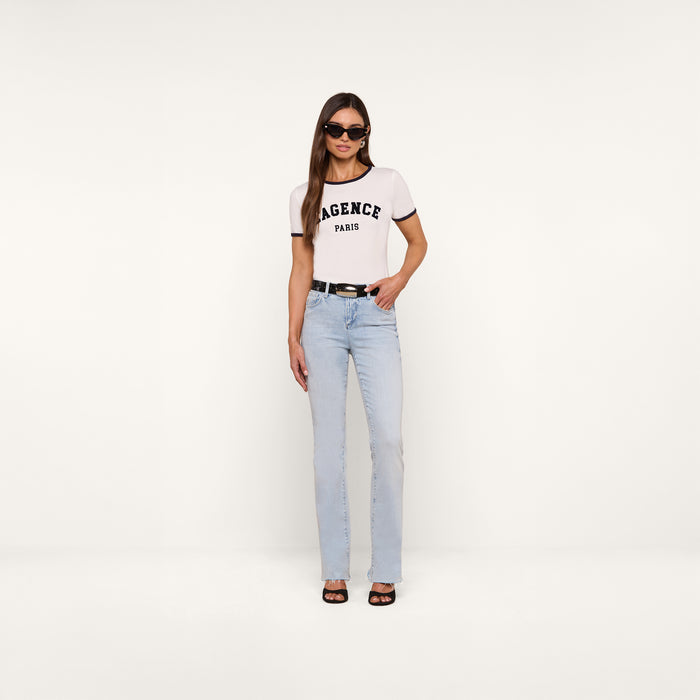 A woman stands against a plain white background wearing sunglasses, light blue jeans with a black belt, black sandals, and the white Tommie Crewneck Tee made from Supima cotton.