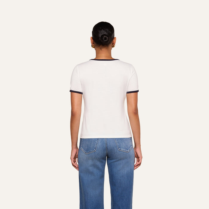 A person stands facing away, wearing the Tommie Crewneck Tee—a white ringer shirt with dark trim—paired with blue jeans. Their hair is pulled back in a bun, and the background is plain off-white.