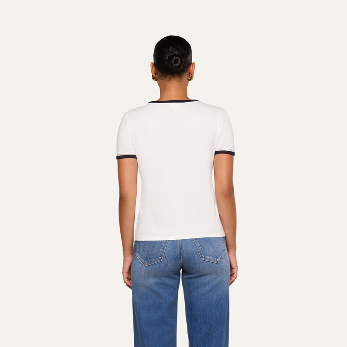 A person stands with their back to the camera wearing the Tommie Crewneck Tee, a relaxed fit white short-sleeve shirt with dark trim, paired with blue jeans against a plain light background.