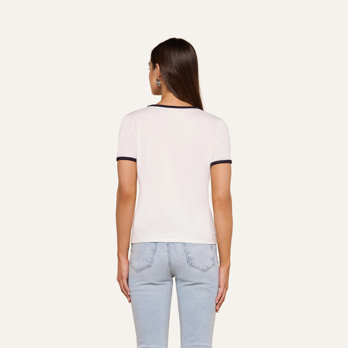 A woman with long brown hair, wearing a relaxed fit Tommie Crewneck Tee in white with black trim and light blue jeans, stands facing away from the camera against a plain background.