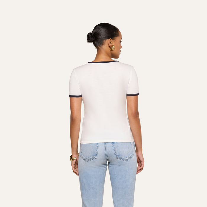 A woman stands facing away, wearing the Tommie Crewneck Tee with dark trim and light blue jeans. Her hair is styled in a neat bun. The background is plain and light.