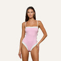 A woman stands against a plain light background, wearing the Tory Bandeau One-Piece Swimsuit—a pink, bouclé fabric one-piece with white trim and thin straps.