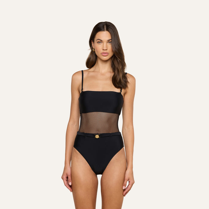 A woman stands against a plain light background, wearing the Tory Bandeau One-Piece Swimsuit — a black one-piece with UPF 50 protection, sheer mesh insets at the midsection, thin straps, and a small gold detail at the waist.