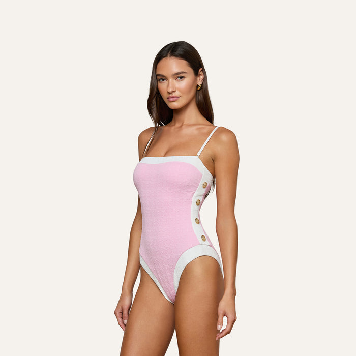A woman models the Tory Bandeau One-Piece Swimsuit, crafted from pink bouclé fabric with white straps and panels, and accented by gold buttons on the side. She stands against a plain light background, gazing confidently at the camera.