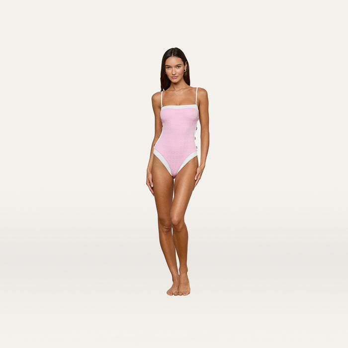 A woman stands barefoot against a plain white background, wearing the Tory Bandeau One-Piece Swimsuit in light pink with thin straps and white trim details, looking directly at the camera with a neutral expression.