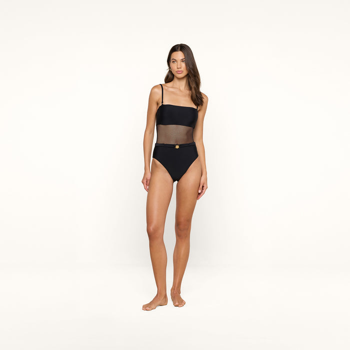 A woman stands barefoot against a white background, wearing the Tory Bandeau One-Piece Swimsuit, which features a one-shoulder design with mesh insets at the waist and high-waisted bottoms.