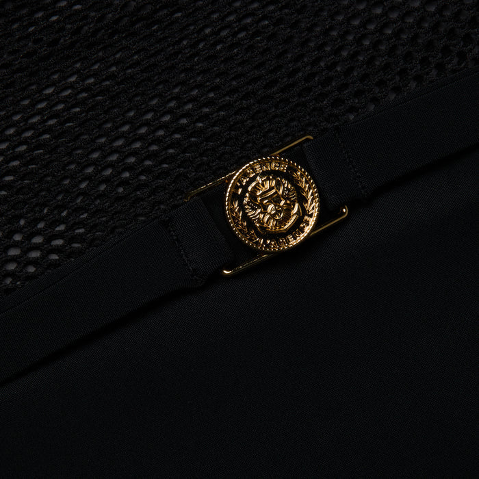 A close-up of a gold Medusa head emblem on the black fabric of the Tory Bandeau One-Piece Swimsuit, shown above a section of black mesh insets.