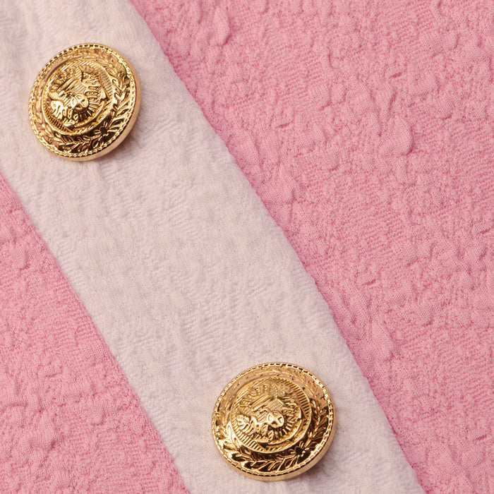 Close-up of two ornate gold buttons on the white bouclé fabric strap of the Tory Bandeau One-Piece Swimsuit, set against a textured pink background.