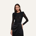 A woman wearing the Trista Bodysuit—a black mesh design with structured shoulder pads—paired with a black satin skirt featuring side draping, stands against a plain light background.