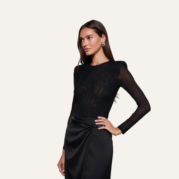 A woman stands against a plain background, wearing the Trista Bodysuit—a black mesh piece with structured shoulder pads—paired with a fitted satin skirt. She has long brown hair, silver earrings, and her hand on her hip.