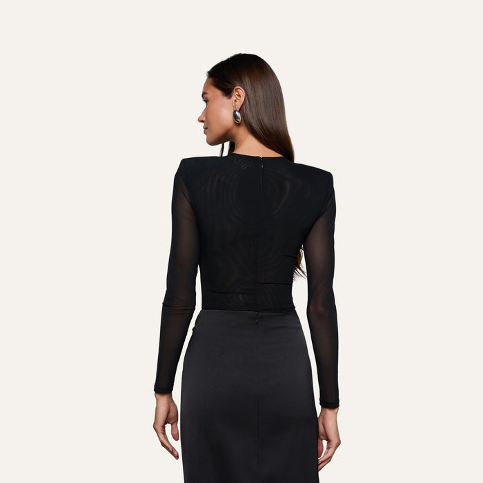 A woman with long brown hair is shown from the back against a plain light background, wearing the Trista Bodysuit—a black mesh, long-sleeve bodysuit—paired with a black skirt.