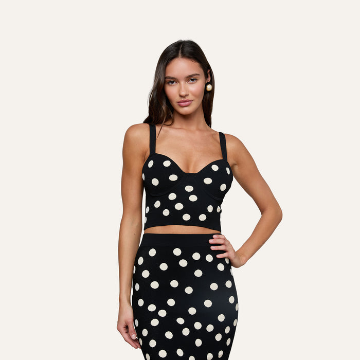 A woman wearing the Tula Knit Bra Top and a matching skirt set with large jacquard polka dots stands with one hand on her hip against an off-white background.