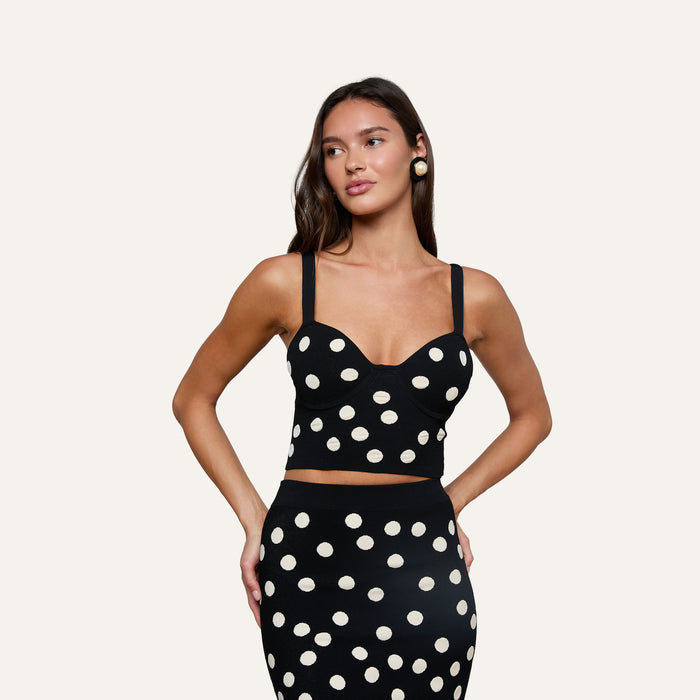 A woman poses with one hand on her hip against a plain light background, wearing the Tula Knit Bra Top—a black compression knit crop top adorned with large white jacquard polka dots—paired with the matching skirt.