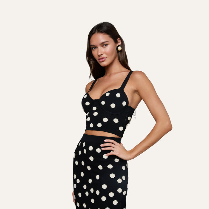 Wearing the Tula Knit Bra Top, a woman poses in a matching black set with large jacquard polka dots and a corset-style design, standing against a plain light background.
