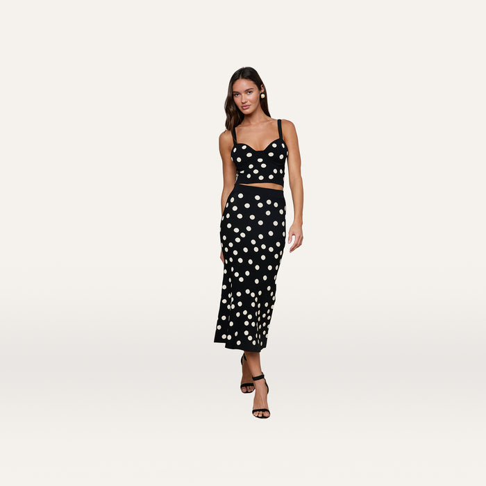 A woman wears the Tula Knit Bra Top, a black corset-style crop top with white jacquard polka dots, paired with a matching midi skirt and black high-heeled sandals, posing against a plain white background.