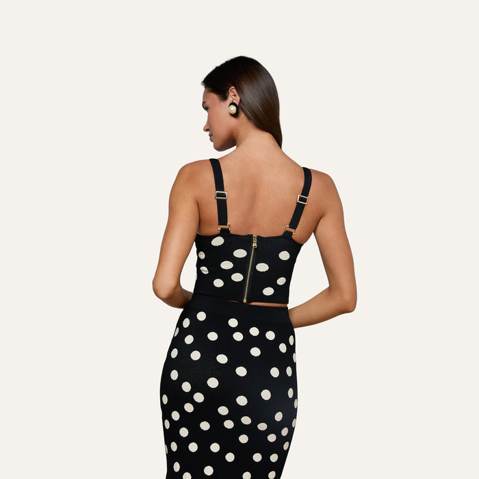 A woman with long brown hair is seen from behind, wearing the Tula Knit Bra Top—a corset-style black crop top paired with a matching skirt featuring large jacquard polka dots. She accessorizes with black earrings and stands against a plain light background.