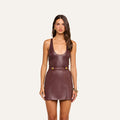 A woman faces forward against a plain light background, wearing the Tullia Scoopneck Tank, a sleeveless mahogany brown vegan leather dress featuring a scoop neck and gold button details at the waist.