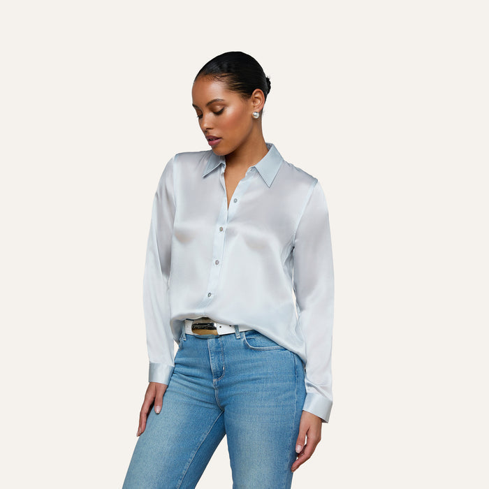 A woman wearing the Tyler Silk Blouse in pale mist blue and blue jeans stands against a plain light background, looking down with one hand resting by her side.