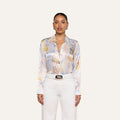 A woman stands against a plain background, wearing the L’AGENCE Tyler Silk Blouse in a pastel pattern, paired with high-waisted white pants and a black-buckled white belt.