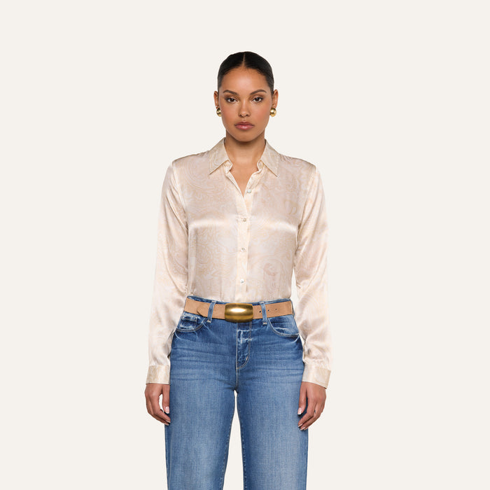 A woman stands facing forward, wearing the Tyler Silk Blouse from L’AGENCE with a subtle paisley print, blue jeans, a gold belt, and gold hoop earrings against a plain light background.