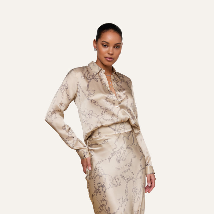 Woman wearing the Tyler Silk Blouse by L’AGENCE—a button-down blouse in a beige chain print—paired with a matching skirt, posing with one hand on her hip against a plain light background.