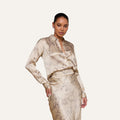 Woman wearing the Tyler Silk Blouse by L’AGENCE—a button-down blouse in a beige chain print—paired with a matching skirt, posing with one hand on her hip against a plain light background.