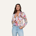 A woman wears the Tyler Silk Blouse—a button-down, long-sleeve pastel blouse featuring a colorful abstract print—paired with light blue jeans, standing against a plain, light background.