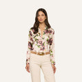 A woman wearing the Tyler Silk Blouse—a long-sleeve, button-down blouse featuring a mauve rose print—paired with cream-colored pants, a tan belt with a gold buckle, and gold earrings, stands against a plain white background.