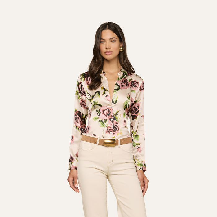 A woman wearing the Tyler Silk Blouse—a long-sleeve, button-down blouse featuring a mauve rose print—paired with cream-colored pants, a tan belt with a gold buckle, and gold earrings, stands against a plain white background.