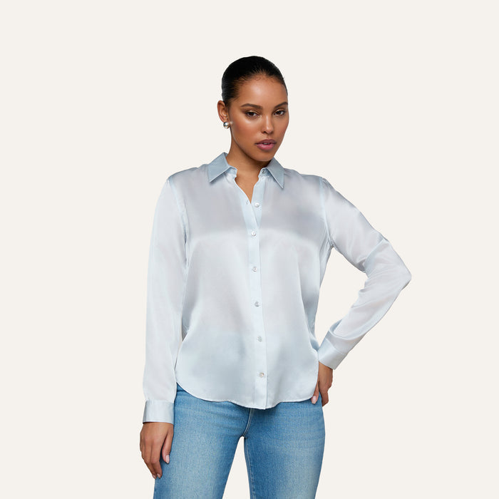 Wearing the Tyler Silk Blouse in pale mist blue and a pair of blue jeans, a person poses against a plain light background with one hand on their hip and the other relaxed by their side.