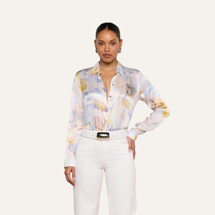 A woman stands facing forward, wearing the pastel patterned Tyler Silk Blouse by L’AGENCE, tucked into high-waisted white pants with a wide belt. Her hair is pulled back, and she accessorizes with earrings against a plain, light-colored background.