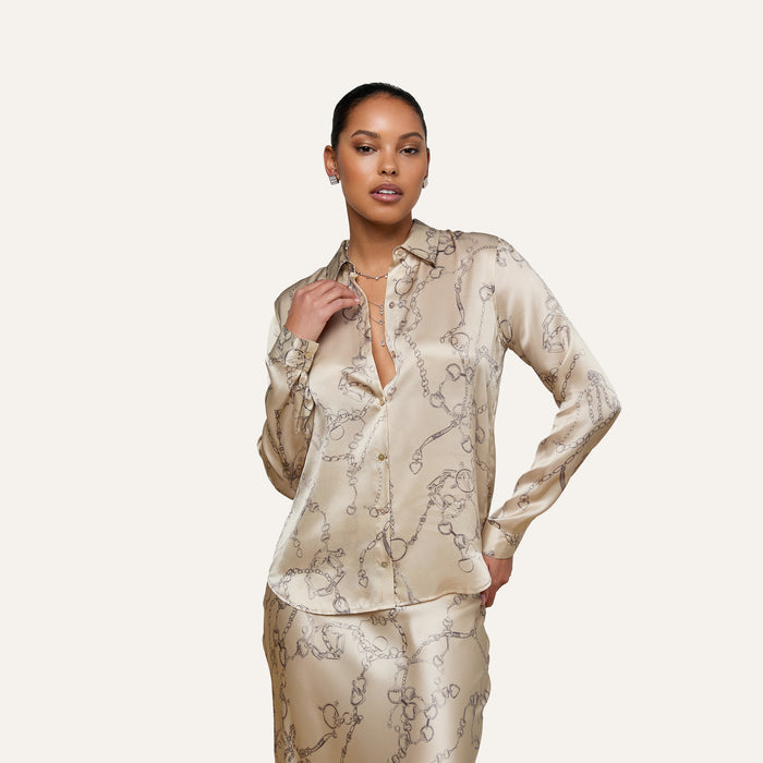 A woman stands confidently against a plain light background, wearing the Tyler Silk Blouse in cream and a matching skirt with a subtle floral pattern; she has one hand on her hip and the other holding her collar.