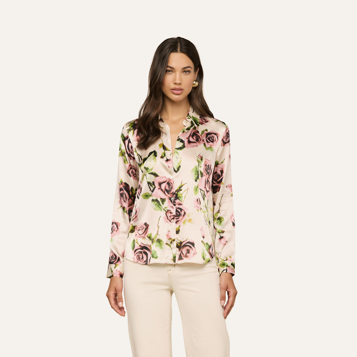 A woman wears the Tyler Silk Blouse—a long-sleeve, button-down silk top featuring a mauve rose print—paired with light-colored pants and stands against a plain, light background.
