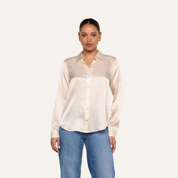 A woman wearing the Tyler Silk Blouse in light beige with a subtle paisley print, paired with blue jeans, stands against a plain off-white background.