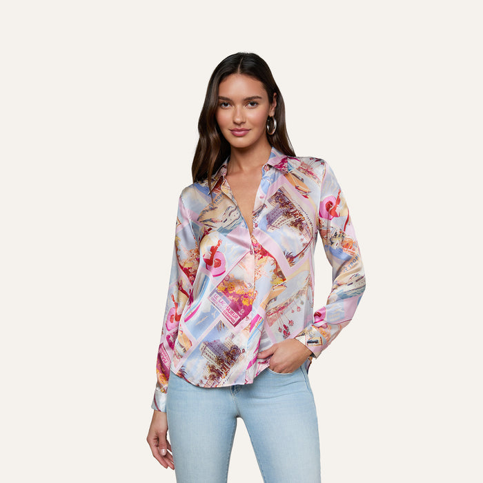 A woman stands and smiles against a light background, wearing the Tyler Silk Blouse with a vibrant pink postcard print and light blue jeans.