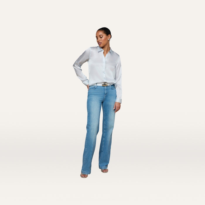 A woman stands against a plain white background, wearing the Tyler Silk Blouse in pale mist blue, tucked into high-waisted light blue jeans with open-toe heels. She has one hand in her pocket and gazes slightly downward to her left.
