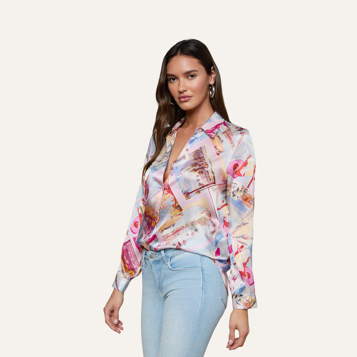A woman stands against a plain, light background, looking at the camera with a slight smile. She is wearing the Tyler Silk Blouse—a colorful, pink postcard print long-sleeve silk button-down—paired with light blue jeans.