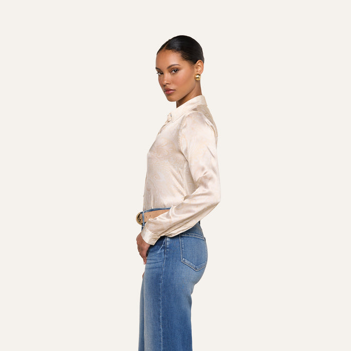 A woman stands in profile against a plain background, wearing the beige Tyler Silk Blouse with long sleeves, blue high-waisted jeans, and gold earrings. Her hair is styled in a neat bun.