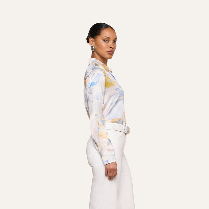 Woman standing sideways, wearing a L’AGENCE Tyler Silk Blouse in pastel colors with long sleeves, paired with white high-waisted pants and a white belt, against a plain light background.