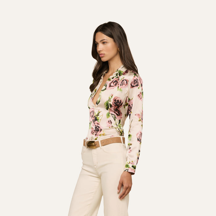 A woman stands in profile wearing the Tyler Silk Blouse, featuring a mauve rose print, long sleeves, and a deep neckline. She pairs it with beige pants and a tan belt. Her long brown hair frames her face, which holds a neutral expression against a plain light background.