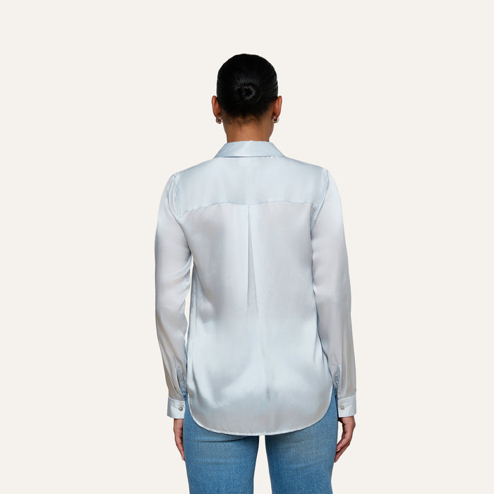 A person with dark hair in a bun wears the Tyler Silk Blouse in pale mist blue and blue jeans, standing with their back to the camera against a plain light background.